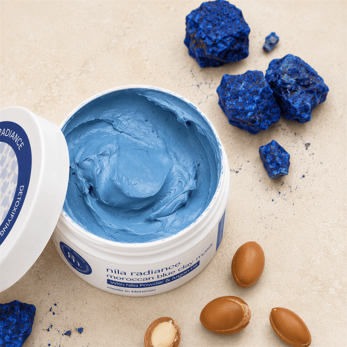 Open white jar of blue nila radiance moroccan blue clay mask beside blue stones and brown nuts on a tan surface.