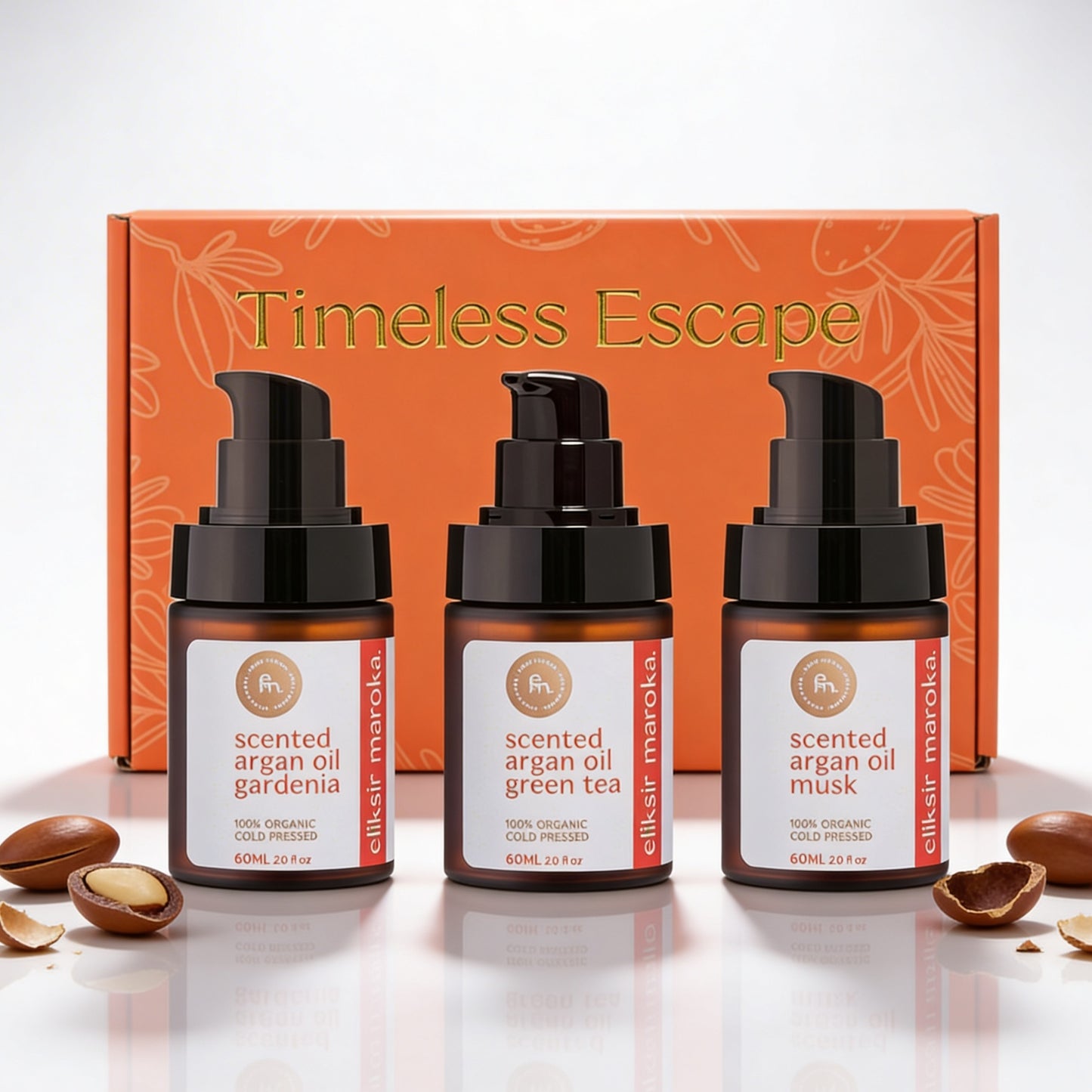 Zestaw prezentowy Timeless Escape od EliksirMaroka. Trzy organiczne, perfumowane olejki arganowe tłoczone na zimno – gardenia, zielona herbata i piżmo – wyeksponowane na tle eleganckiego pomarańczowego pudełka i autentycznych orzechów arganowych. | Timeless Escape gift set by EliksirMaroka. Three organic, cold-pressed scented argan oils – gardenia, green tea, and musk – displayed against an elegant orange box and authentic argan nuts.