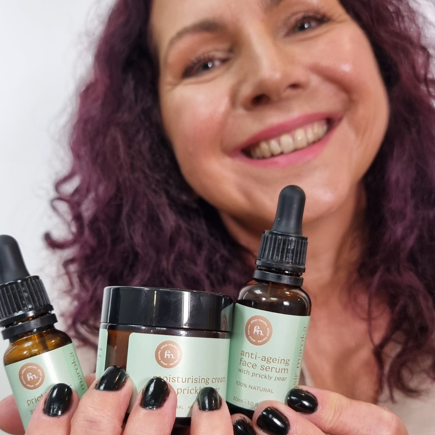 Uśmiechnięta klientka polecająca z bliska kompletny zestaw z opuncją figową EliksirMaroka: 100% naturalne serum przeciwzmarszczkowe, krem nawilżający i olejek. | Smiling customer recommending up close the complete EliksirMaroka prickly pear set: 100% natural anti-ageing serum, moisturising cream, and oil.